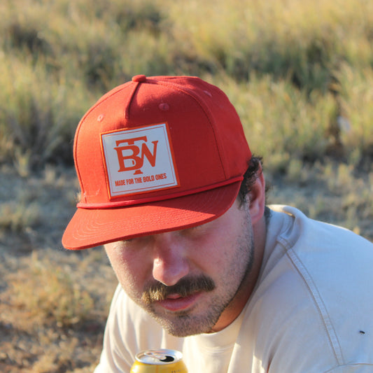Man wearing an orange cap with a logo, holding a can, in a desert-like setting